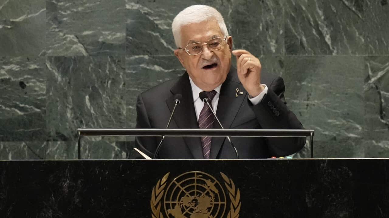 A man gestures with his right hand as he speaks into two microphones on a podium with a United Nations logo in a large, marbled hall. The man is wearing a suit and glasses and has white hair.