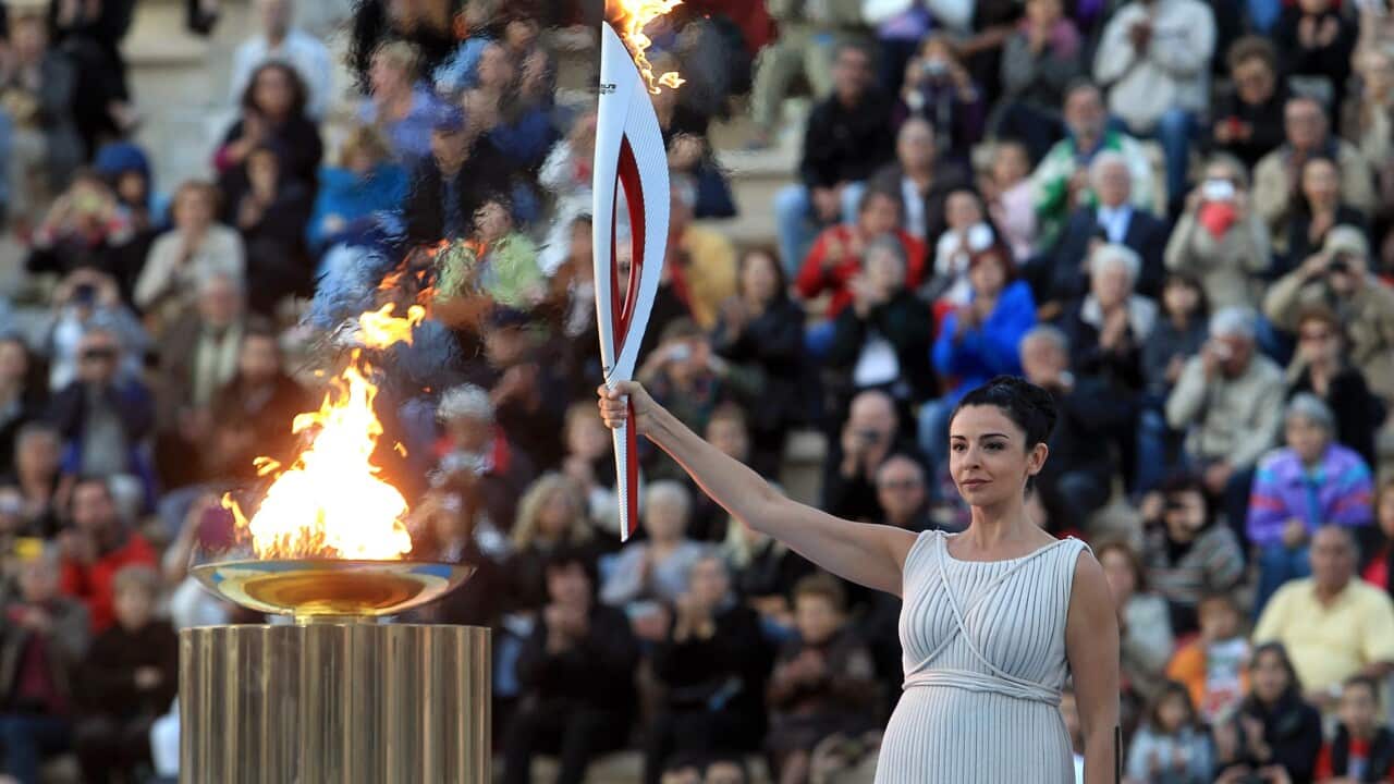 a woman in traditional ancient greek clothing lights a flame on a greek column with a torch.