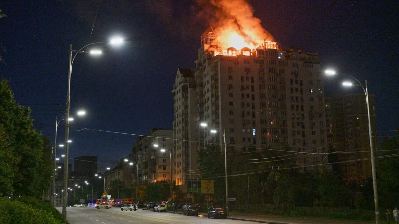 A high-rise residential building's upper floors and penthouse are engulfed in large, bright orange flames and heavy smoke at night, viewed from an adjacent street lined with lit lampposts.