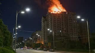 A high-rise residential building's upper floors and penthouse are engulfed in large, bright orange flames and heavy smoke at night, viewed from an adjacent street lined with lit lampposts.