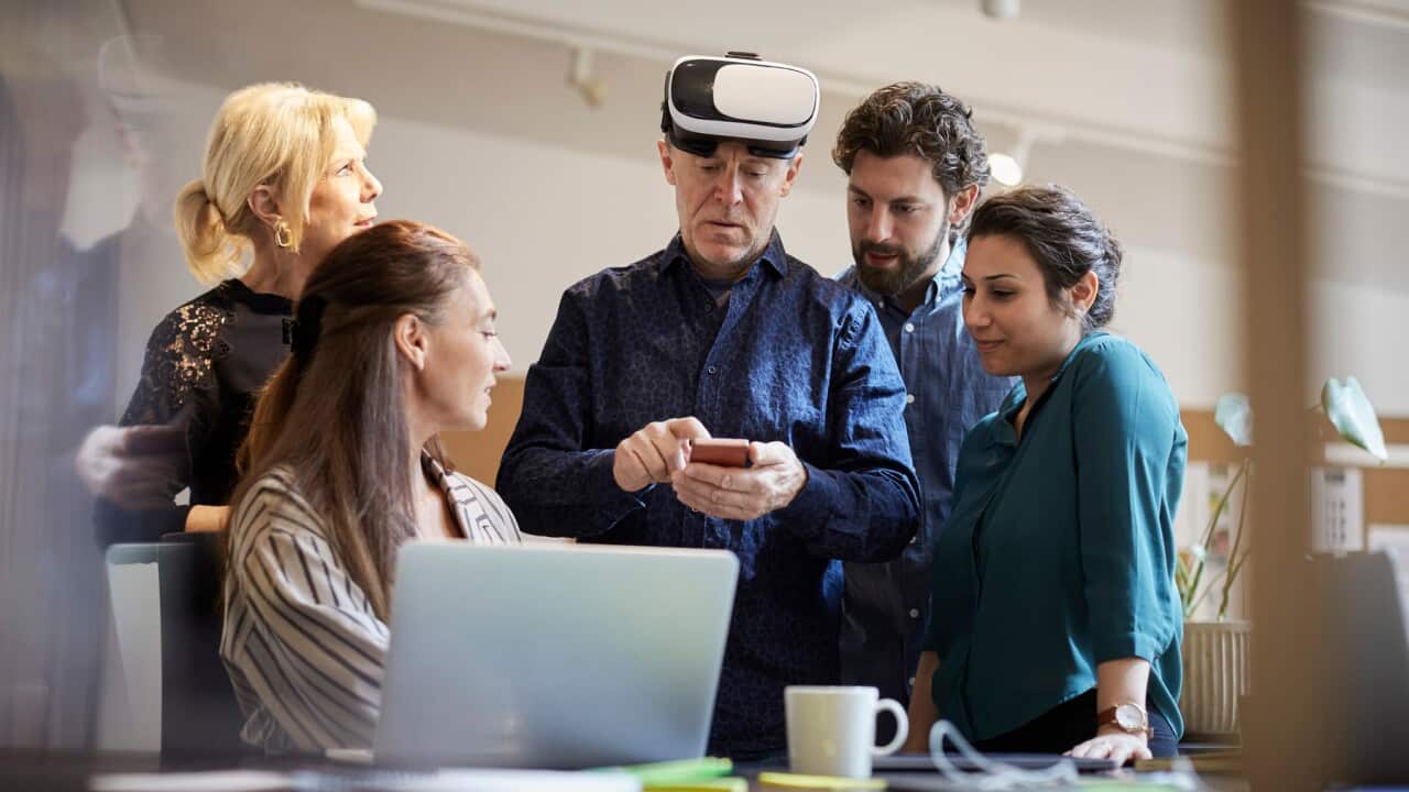 Mature businessman with virtual reality simulator using smart phone while standing amidst colleagues