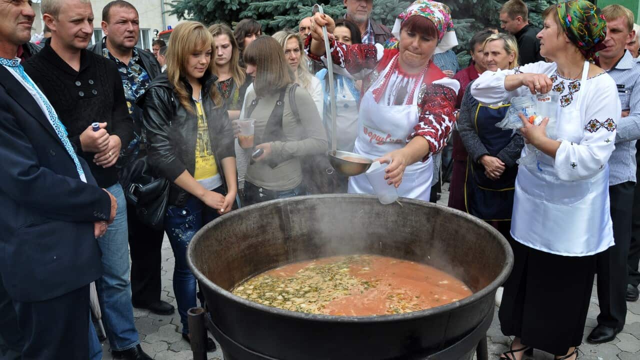 A woman ladles a portion of borsch into a cup out of a large pot.
