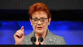 A woman with red hair, wearing glasses and a plaid blazer looks up. Her hand is raised as she speaks.