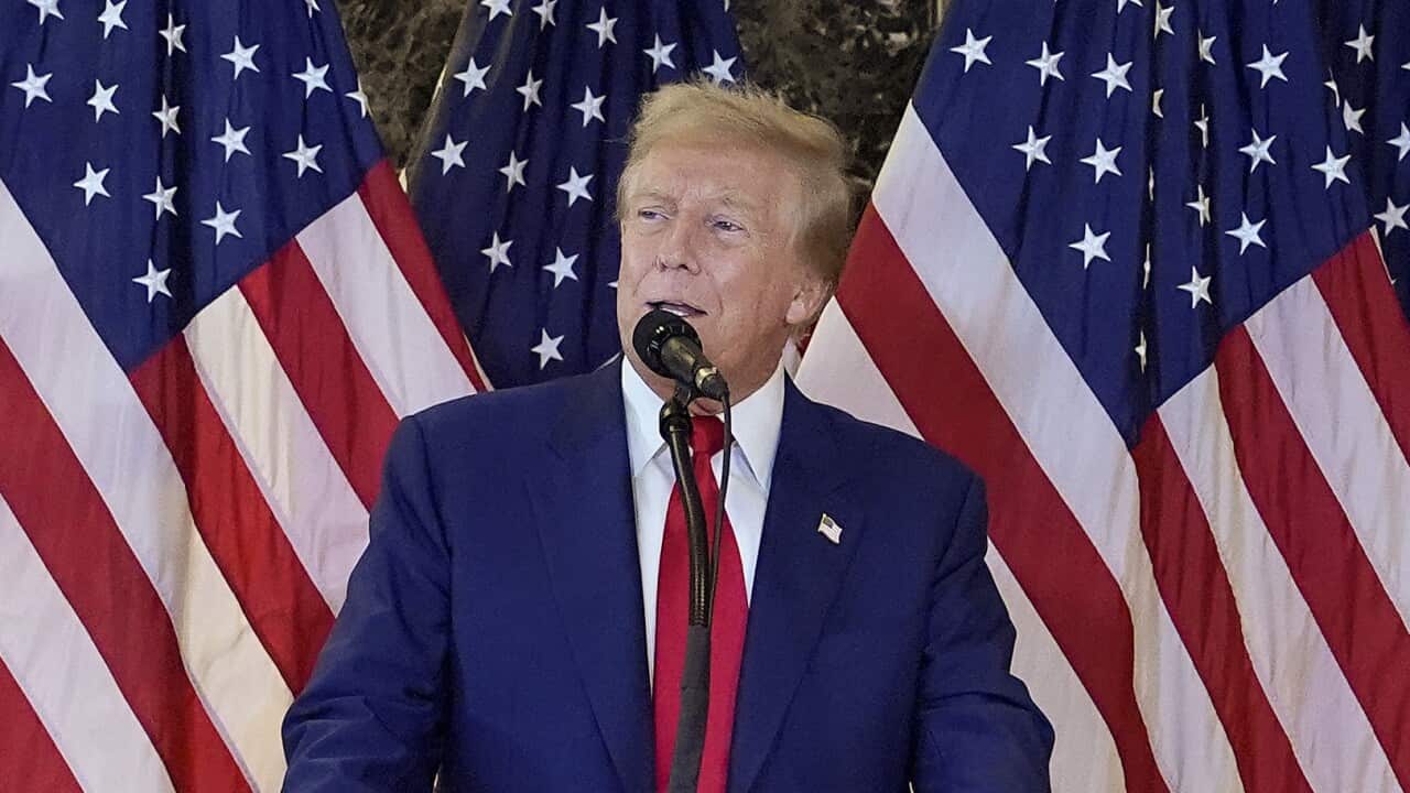 Donald Trump speaking into a microphone in front of a row of American flags.