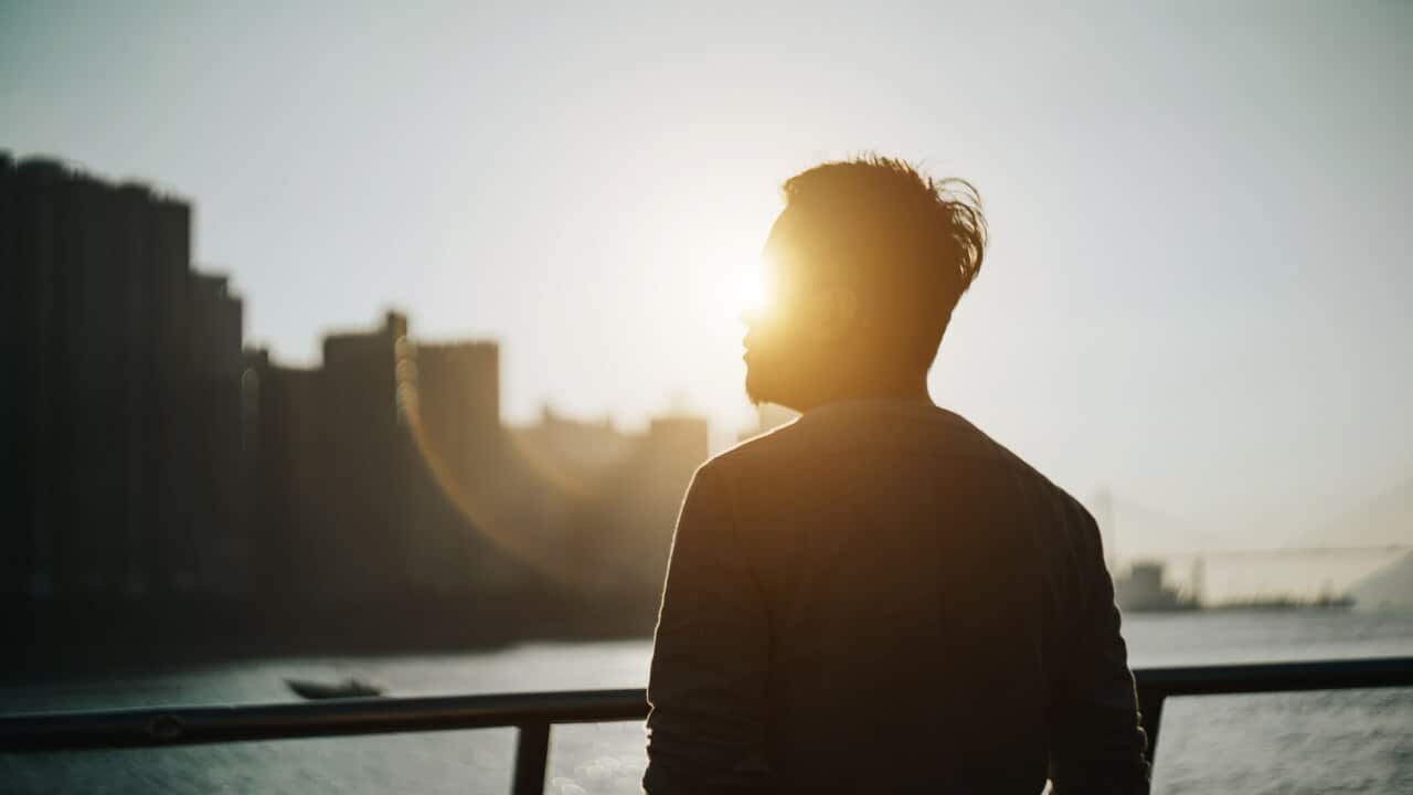 Silhouette of young man standing against urban cityscape and harbour looking up to sky in deep thought