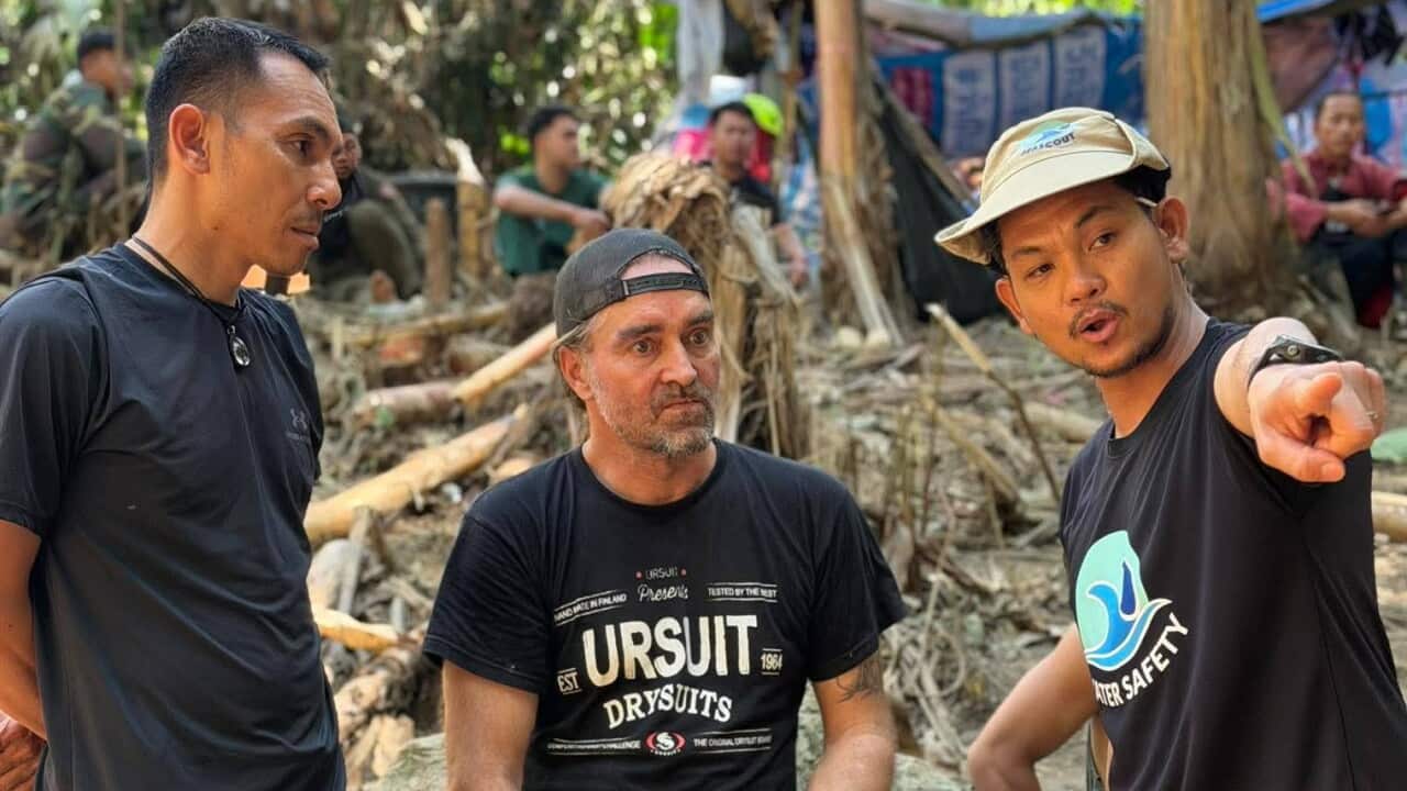 Three men stand in a wooded area. One man is pointing as the other two look on.