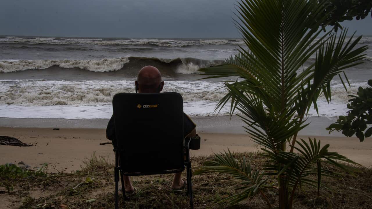A tourist from Perth watches Tropical Cyclone Narelle roll into Port Douglas over the Coral Sea. The man is photographed with his back to the camera, sitting on a camp chair in the grassy dunes of a sodden beach. Beyond him is a grey sky and brown sea that heralds the cyclone's arrival.