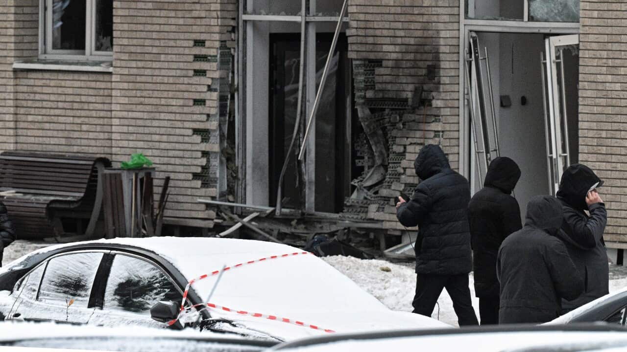 Site of an explosion with four people besides a blown off wall and a car covered in snow.