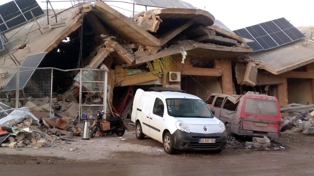 A collapsed building with large concrete slabs and solar panels among the debris. The structure is heavily damaged, with twisted metal and rubble scattered. A white van and a red vehicle are parked in front of the wreckage, partially covered in dust and debris.
