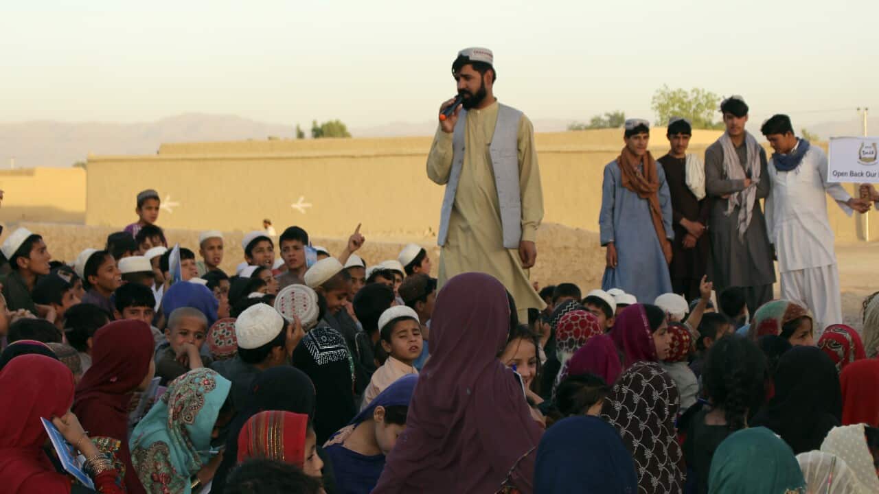 A man speaking into a microphone while a group of children sit down near him