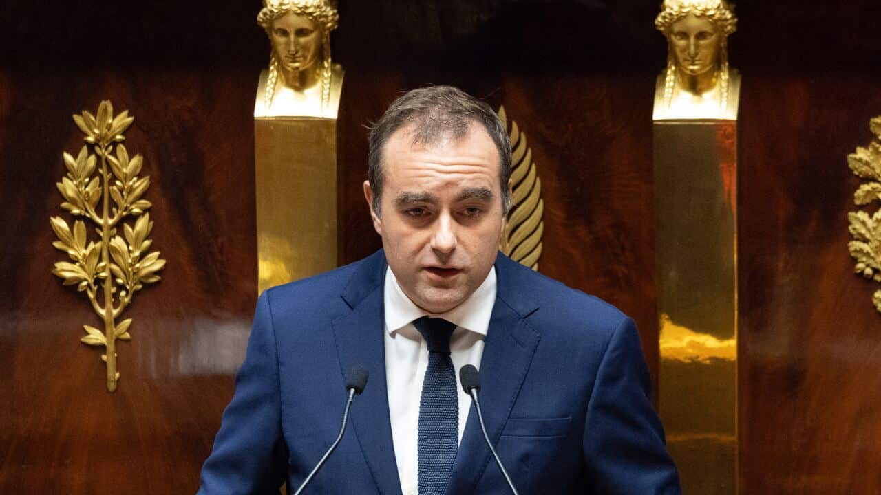 A man in a blue suit stands at a wooden podium holding a folded piece of paper.