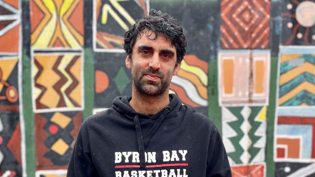 A man in a black hoodie that reads "Byron Bay Basketball" standing in front of a colourful wall.