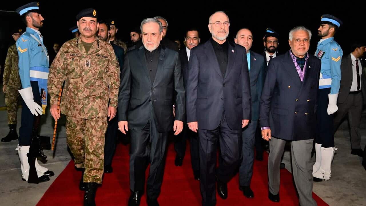 One person in a military uniform and three other people wearing suits are walking on a red carpet. Two soldiers wearing blue uniforms and other people in their entourage are behind them