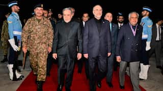 One person in a military uniform and three other people wearing suits are walking on a red carpet. Two soldiers wearing blue uniforms and other people in their entourage are behind them