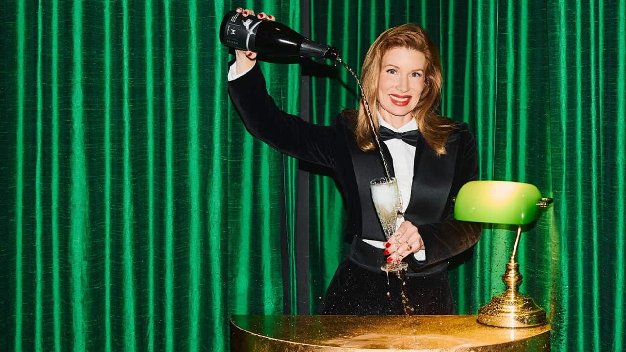 A woman pouring champagne into a glass behind a gold podium with a gold lamp on it. She is in front of a bright green curtain.
