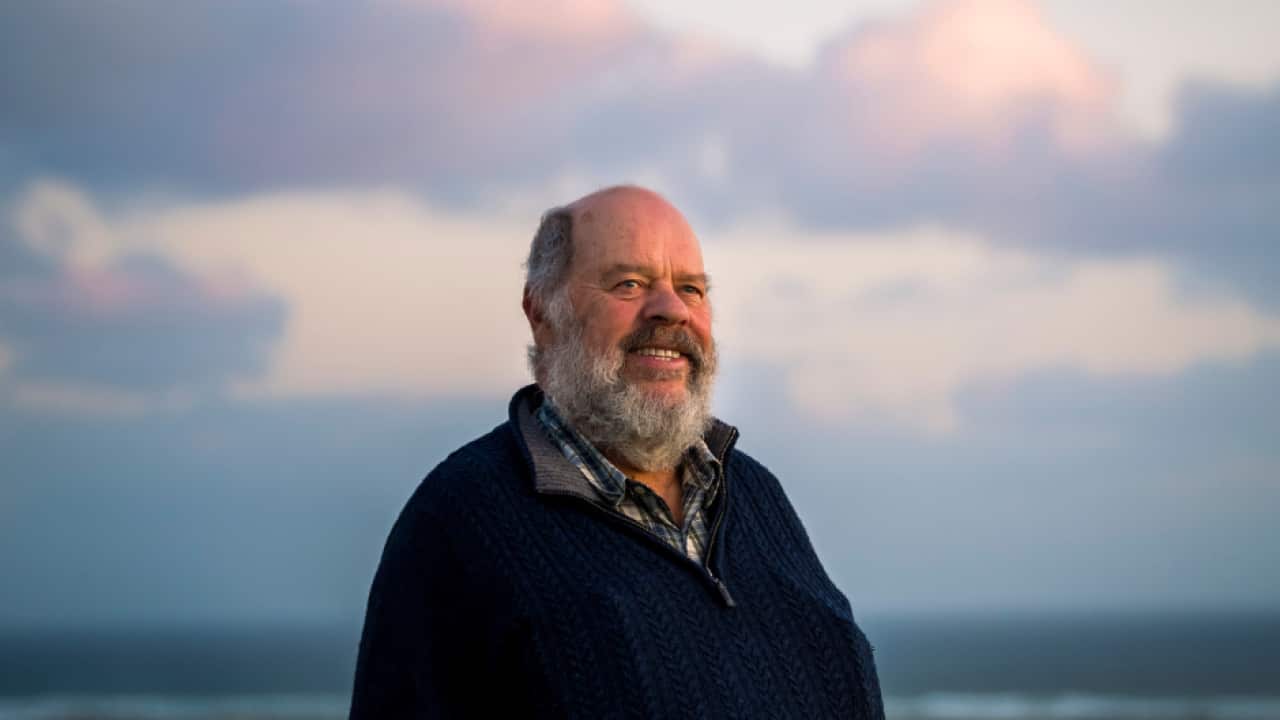 Denis Rose, a Gunditjmara Traditional Owner in his 70s, wearing a navy fleece and standing near the ocean at dusk.