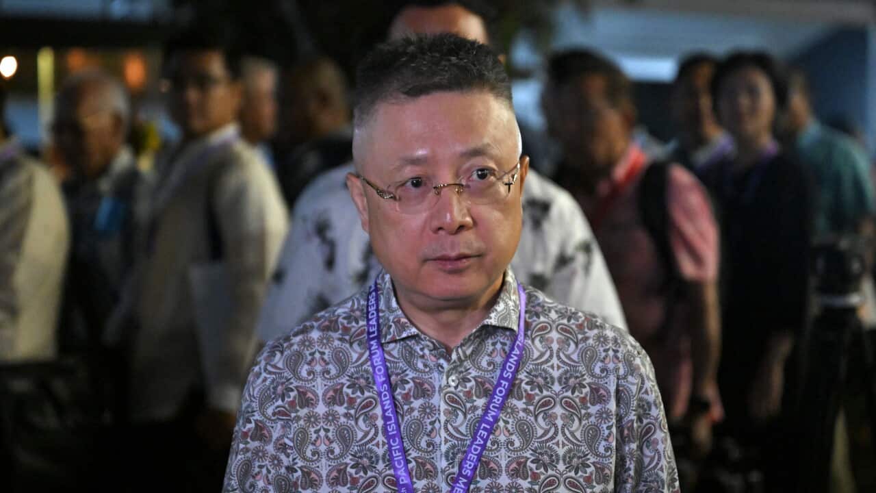 A man wearing glasses and a patterned shirt with a conference lanyard stands among a crowd at an indoor event, with other attendees blurred in the background.