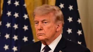 Trump in a navy suit standing in front of an American flag with a serious expression.