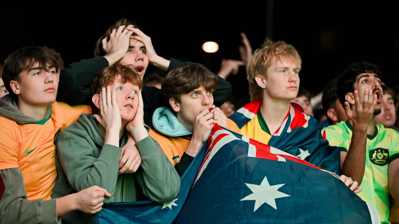 A group of young sports fans, some wearing Australian national colours and holding an Australian flag, display expressions of shock, worry, and disbelief at an outdoor event at night.