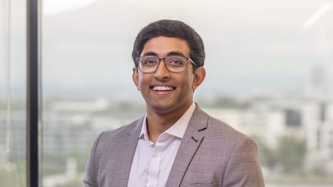 a close up of a young man in a suit and glasses smiling in a glass office