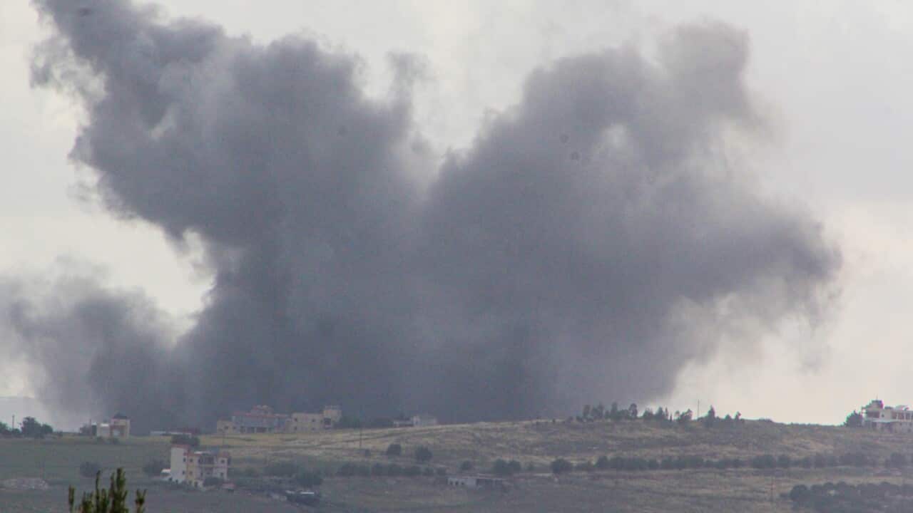 A large plume of dark smoke rises into the sky above an open landscape with scattered buildings and fields.