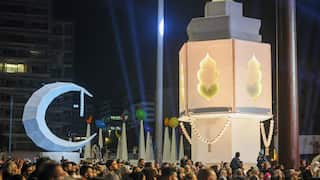 A large crowd of people gathers at night in a public square featuring giant illuminated Ramadan decorations, including a massive white crescent moon and a towering lantern structure. Blue spotlights beam into the sky behind the festive display.
