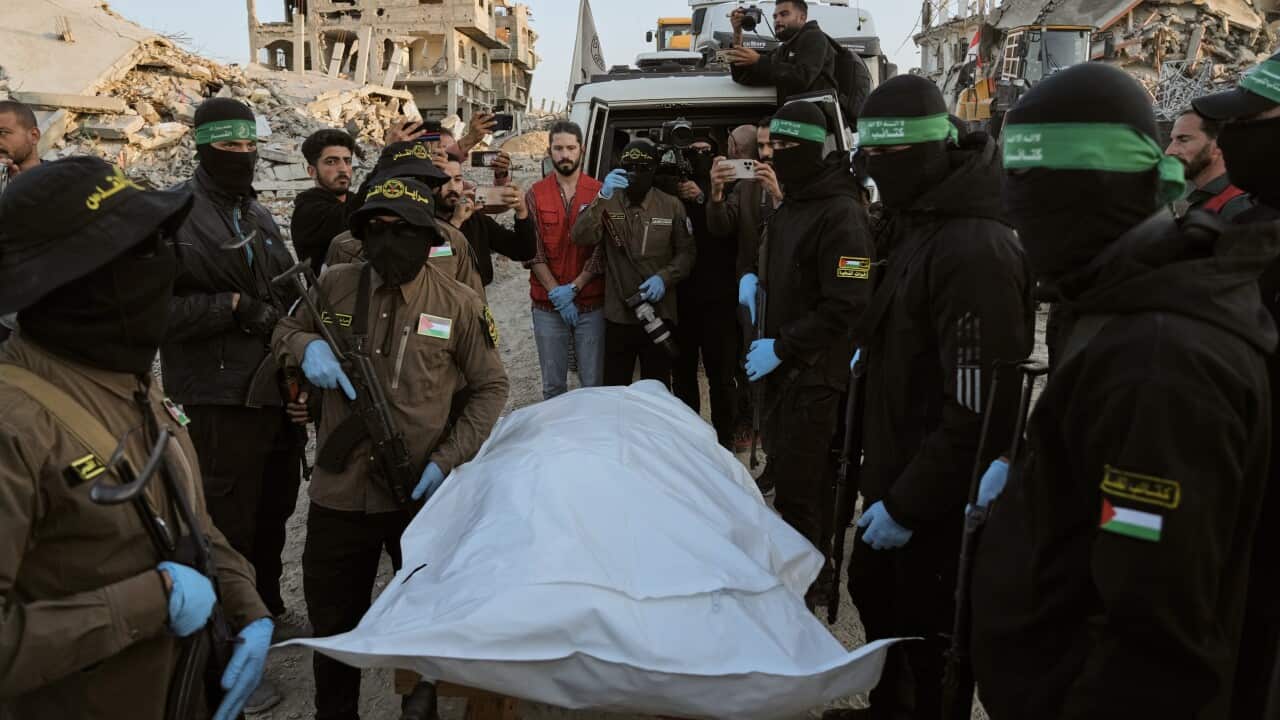 Masked and armed men stand around covered human remains.