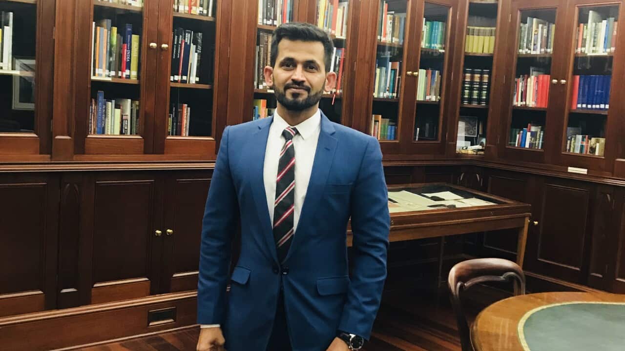 A man wearing a suit stands in a room featuring walls lined with books in wooden bookcases and glass doors