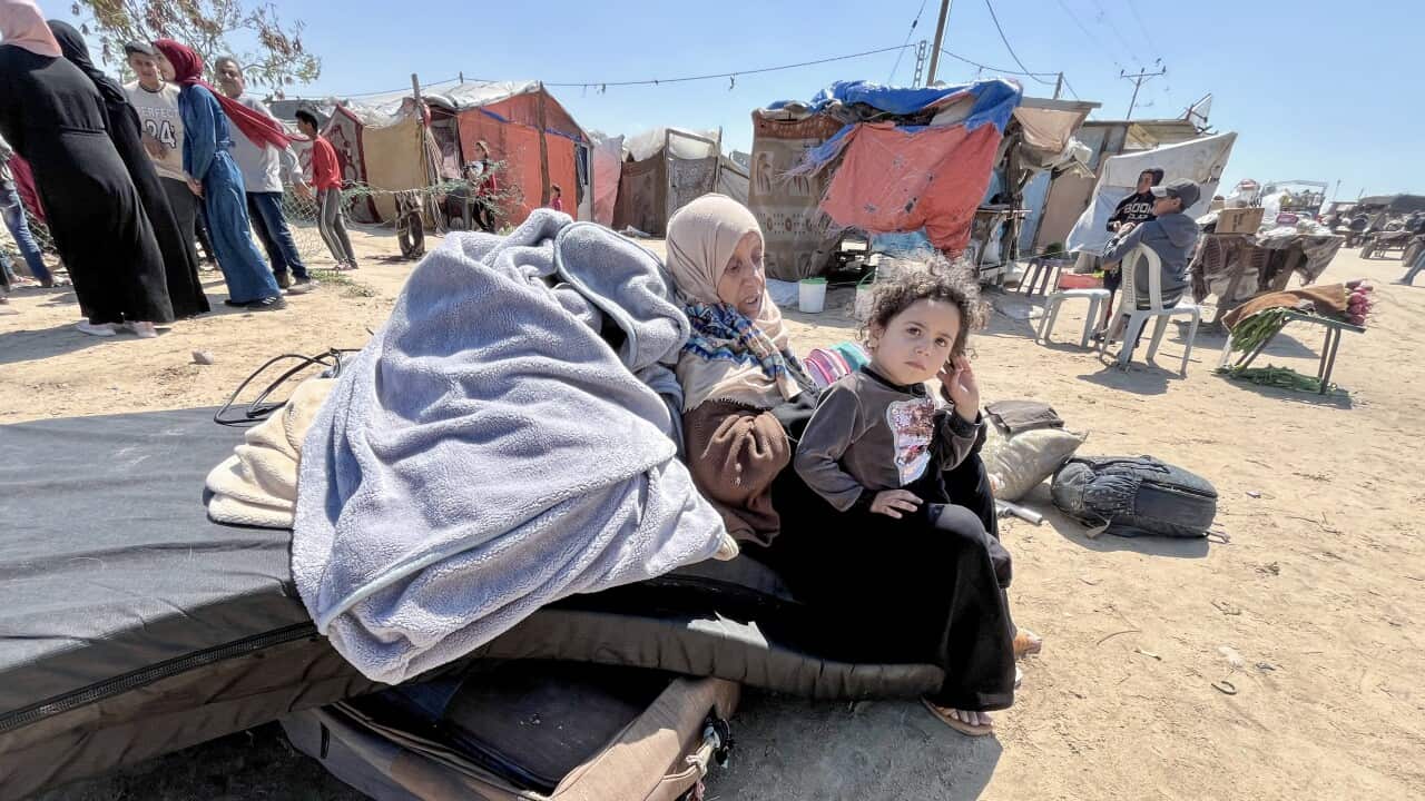 A child sits on their mother's lap surrounded by their belongings with makeshift camps for refugees seen in the background.