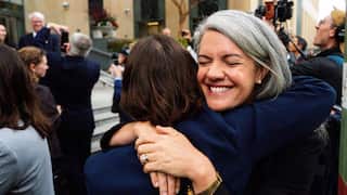 Two women embrace outside a building while a crowd and camera crews gather in the background. One woman with grey hair is facing the camera, while the other has her back to the camera.