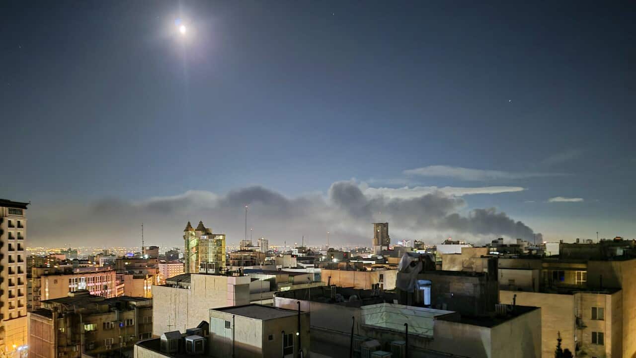 Plumes of thick, dark smoke rise from a distant point on the horizon of a sprawling city skyline at night under a glowing moon.