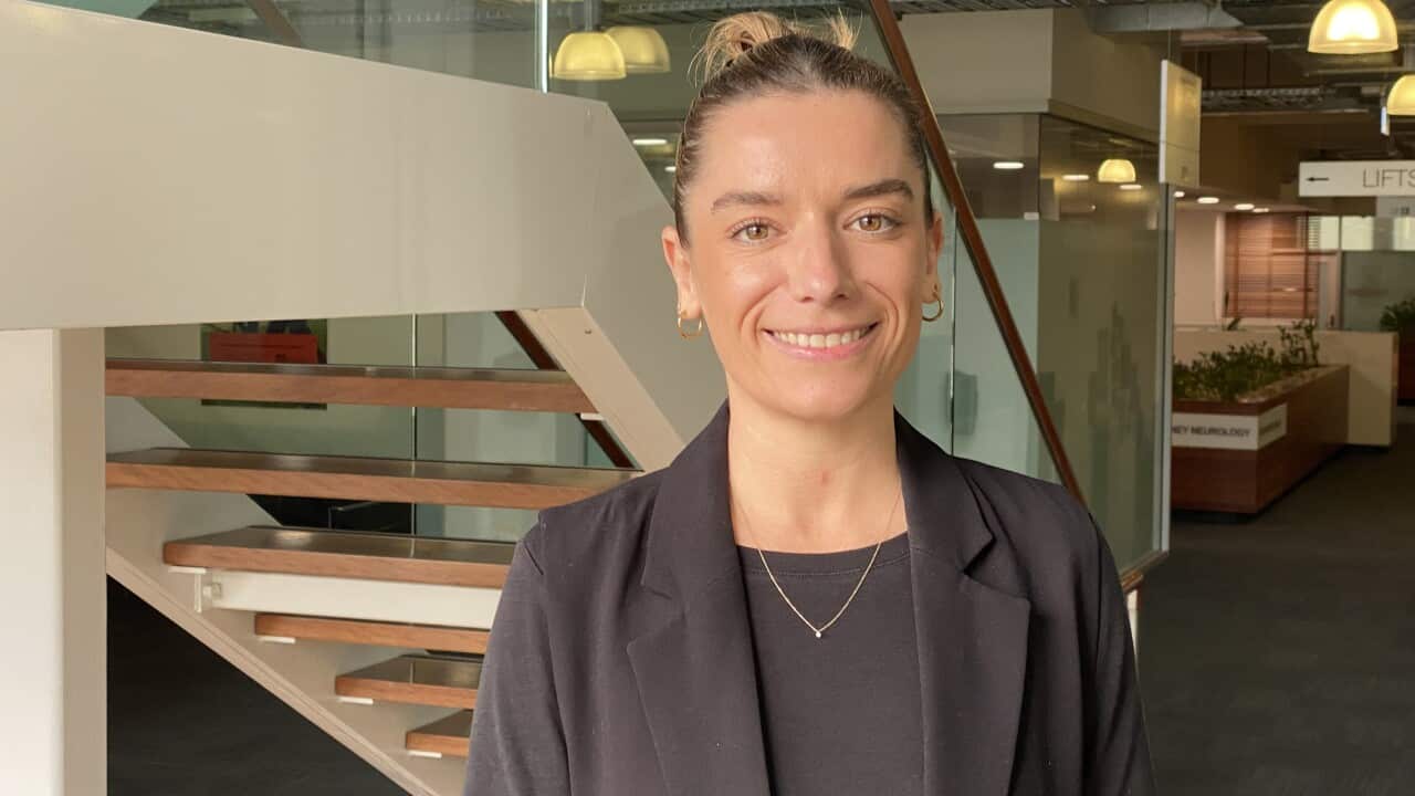A woman in a black blazer and shirt is smiling as she stands in front of a staircase.