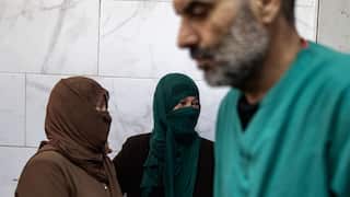 One woman in a brown head covering and another woman in a green head covering console each other. A medic wearing a green uniform is seen walking in front of them