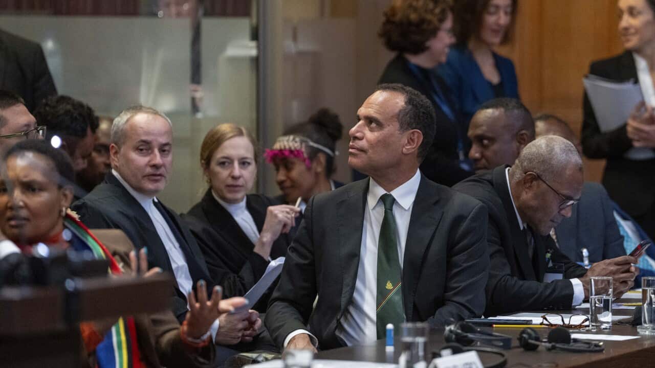 A man in a dark suit with a dark green tie looks behind him, surrounded by people in suits and women in colourful traditional dresses.