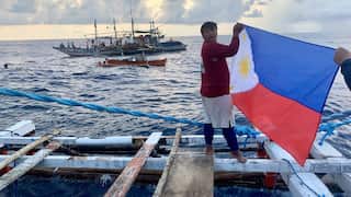 A man in a red shirt stands on a wooden raft at sea. He holds a Philippines flag in his hands. Behind him is a fishing boat and a canoe.
