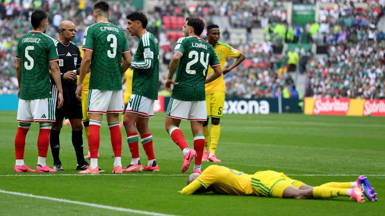 A South African footballer in a yellow jersey lying on the pitch, a group of Mexican players in green speak with a referee in the background