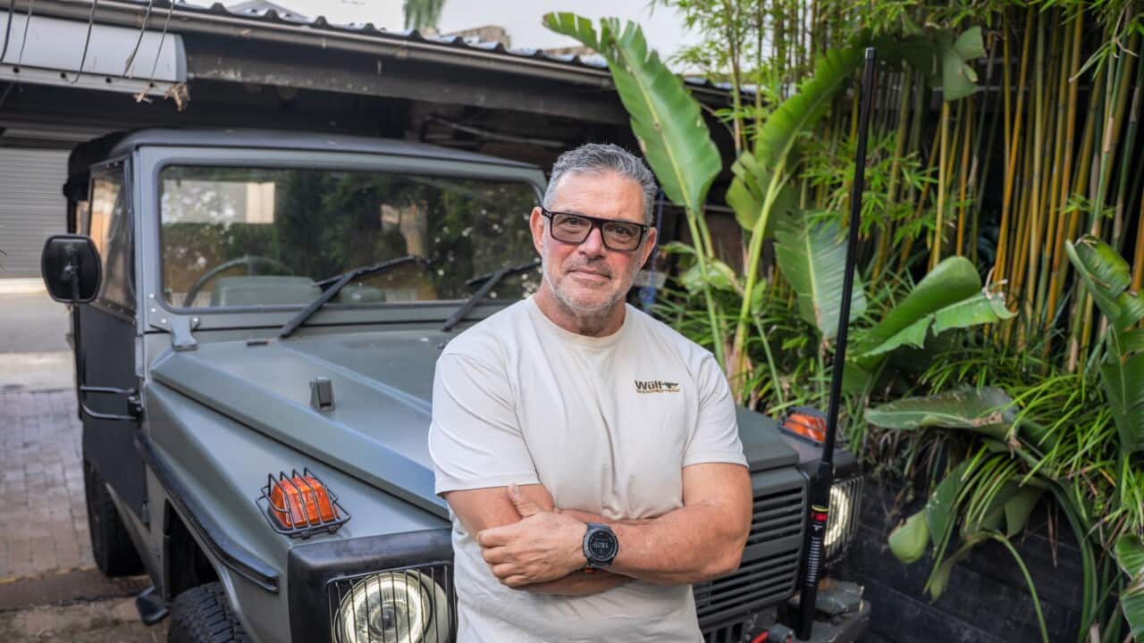 A man in a grey round-neck t-shirt stands in front of his converted military wagon.