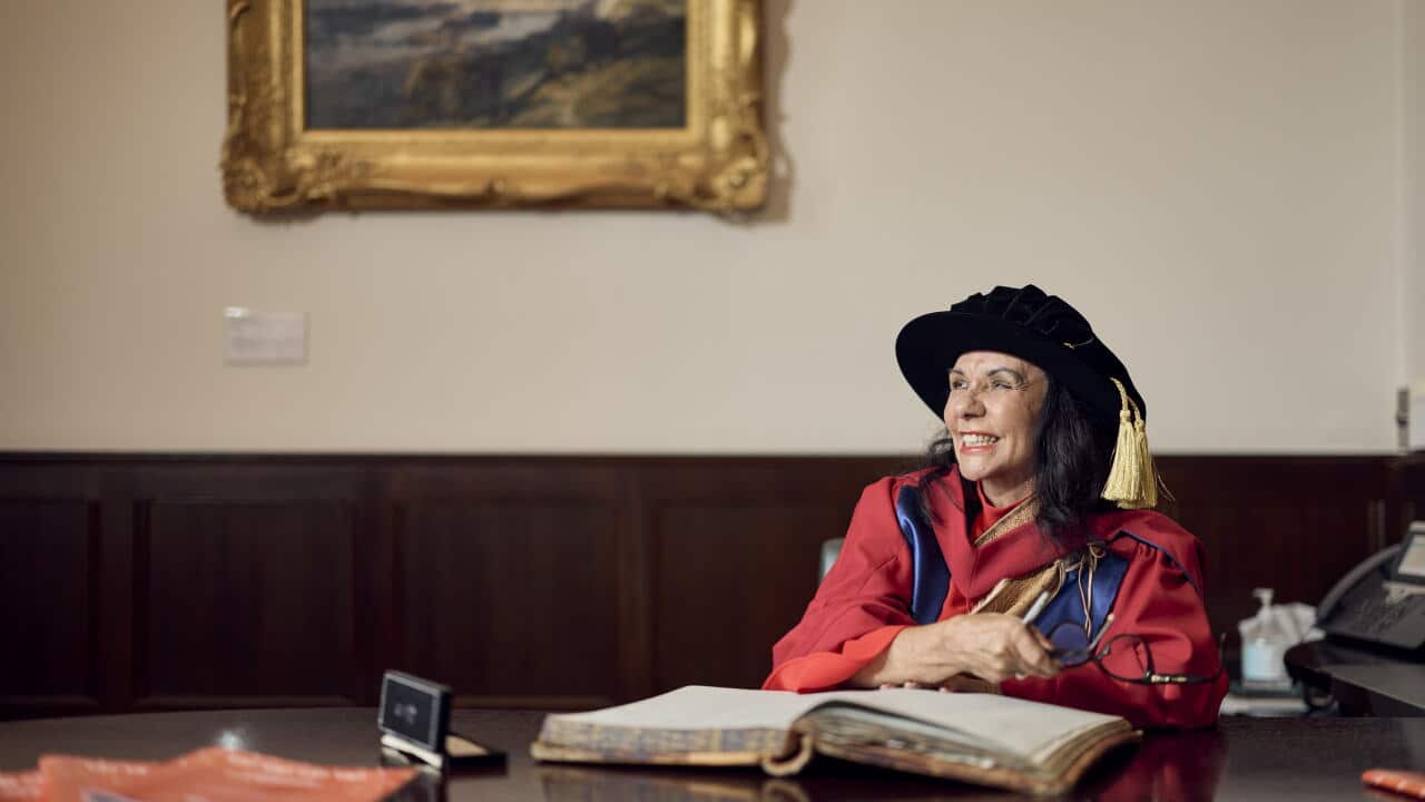 A smiling woman in university regalia, sitting at a large desk.