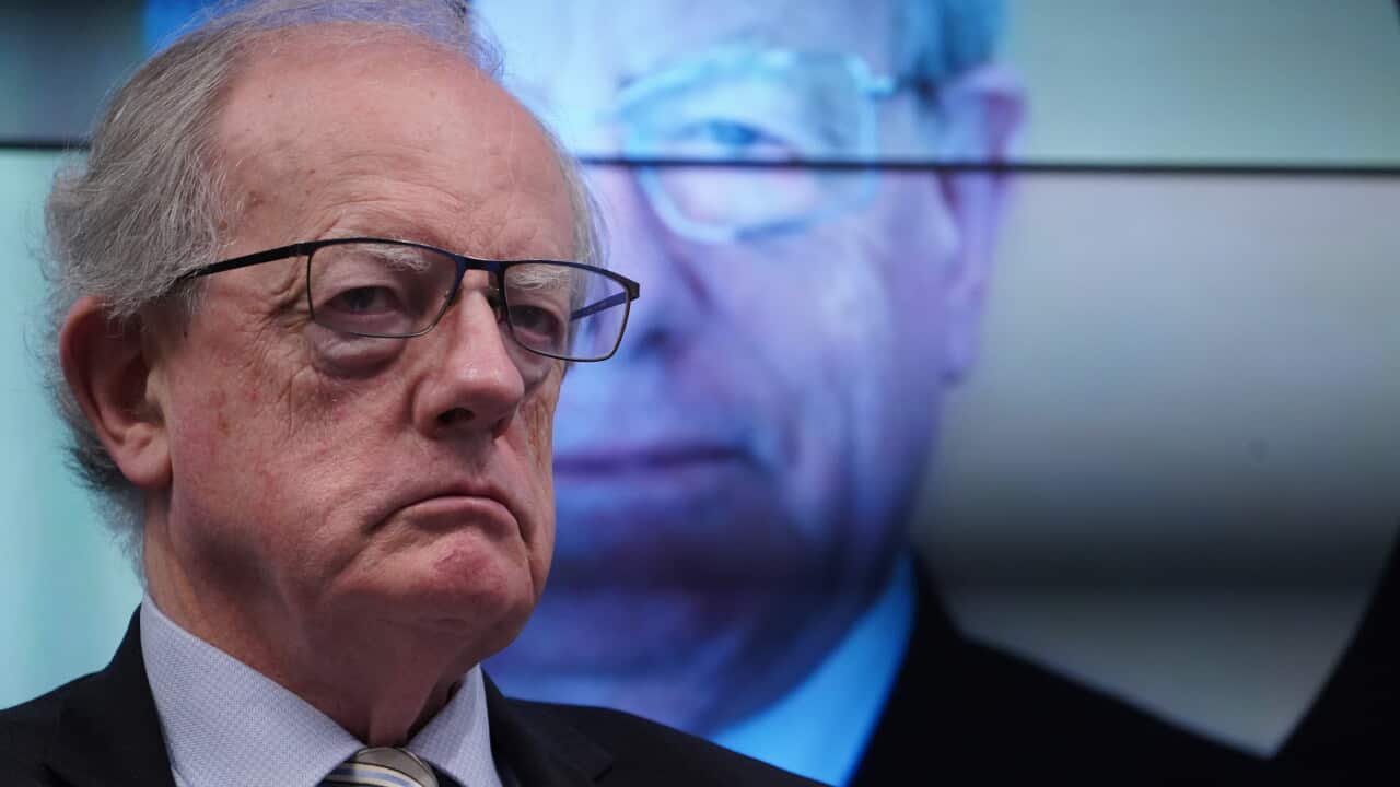 An older man in glasses stands in front of a projected image of himself.