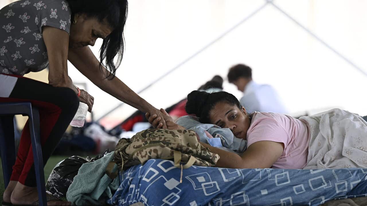 A woman sits in a chair leaning down and holding the hand of a second woman in a makeshift bed.