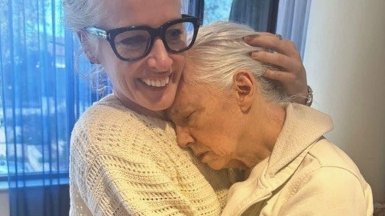 a middle aged woman with thick framed glasses hugs an elderly woman with her eyes and her head on her chest