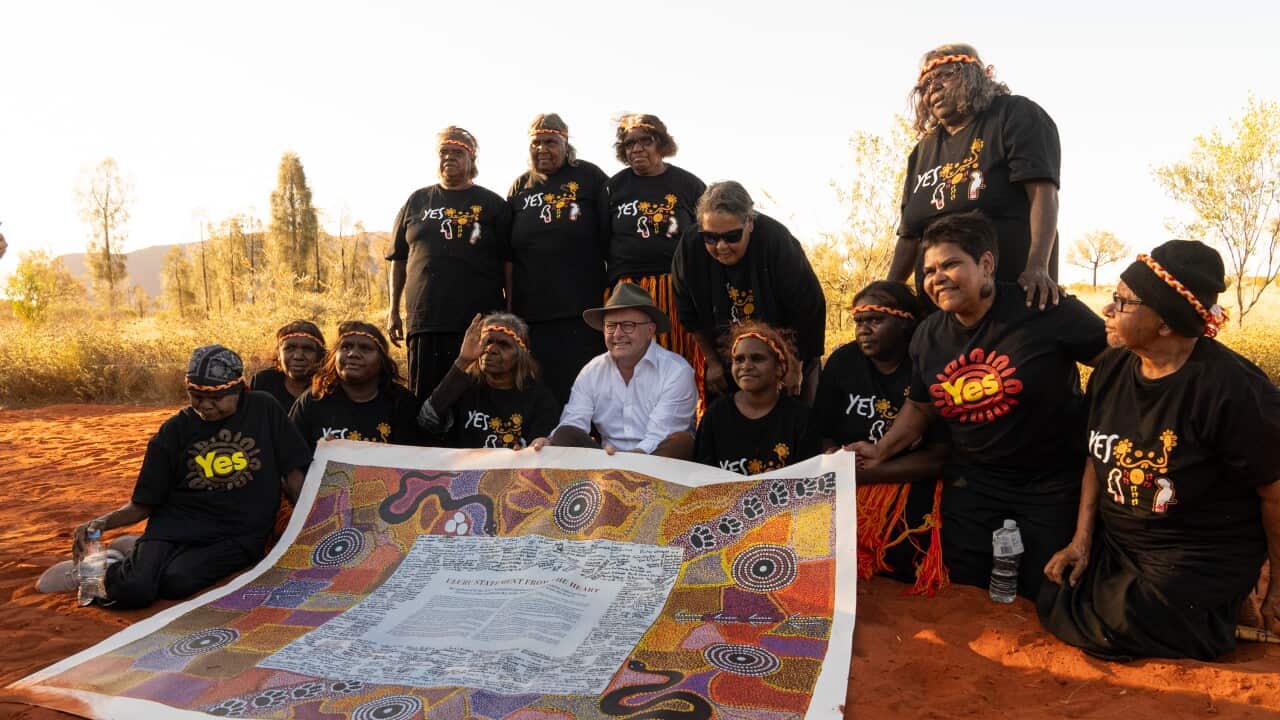 the prime minister in an akubra sits in the red dirt around uluru, visible in the background, with local women sitting around him holding the Uluru statement from the heart.