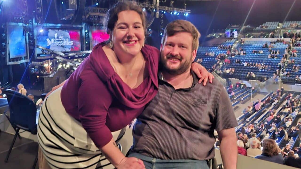 a young couple are at les miserables production, the woman in a purple top smiles and has her hand on her male partner who has a beard and is sitting in a wheelchair