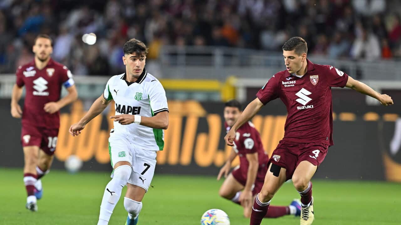 A football player in a maroon jersey runs with a football, with another player in a white and green jersey chasing him. Two other players in maroon jerseys are seen in the background