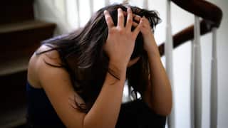 A person sits on a wooden staircase with their head bowed and hands clutching their hair, conveying a sense of distress or overwhelm.