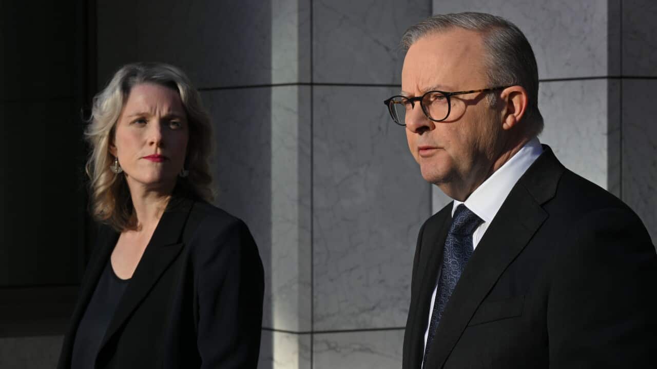 A man wearing a suit, tie and glasses speaks while a woman wearing formal attire listens.