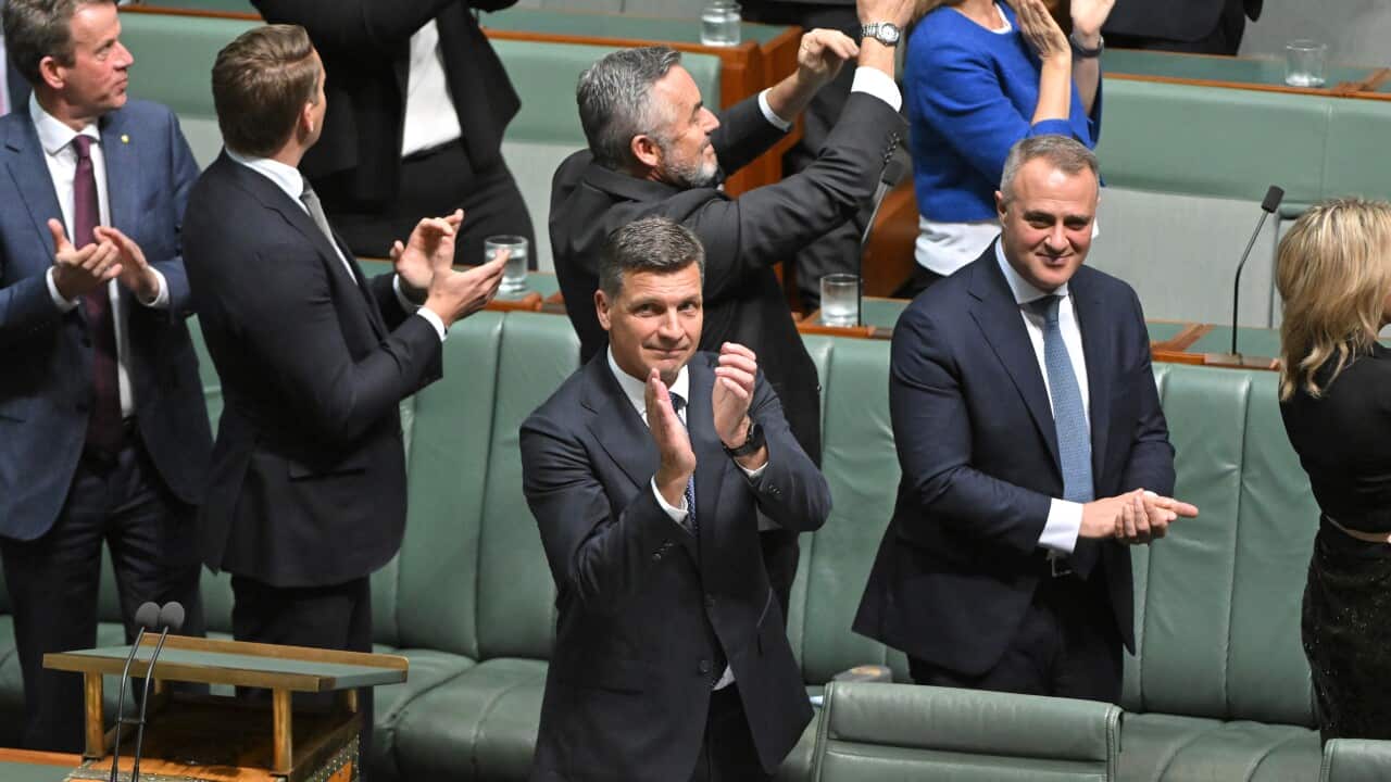 A man in a suit claps his hands and smiles while standing with other in the House of Represenatives.
