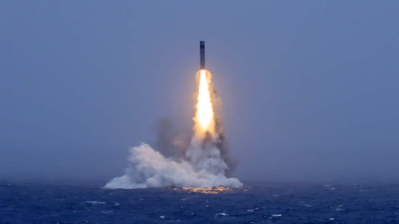 A missile launches vertically from the ocean, creating a bright orange exhaust flame and thick white smoke against a dark, overcast sky.
