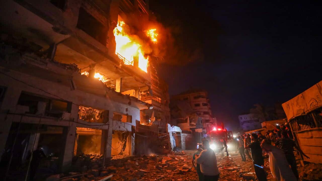 Multi-story building engulfed in flames at night, with debris covering the street below as emergency vehicles and bystanders gather nearby.
