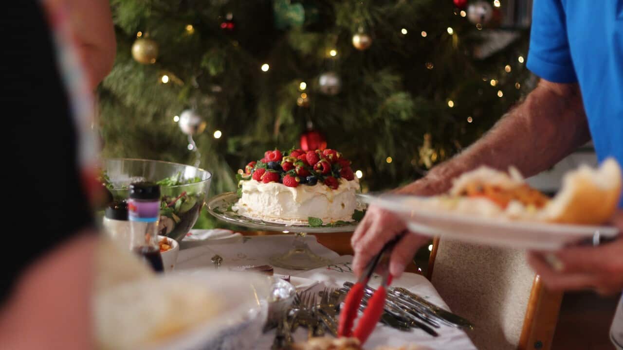 A pavlova on a table with a Christmas tree in the background. A person wearing a blue shirt is piling his plate with food in the foreground.
